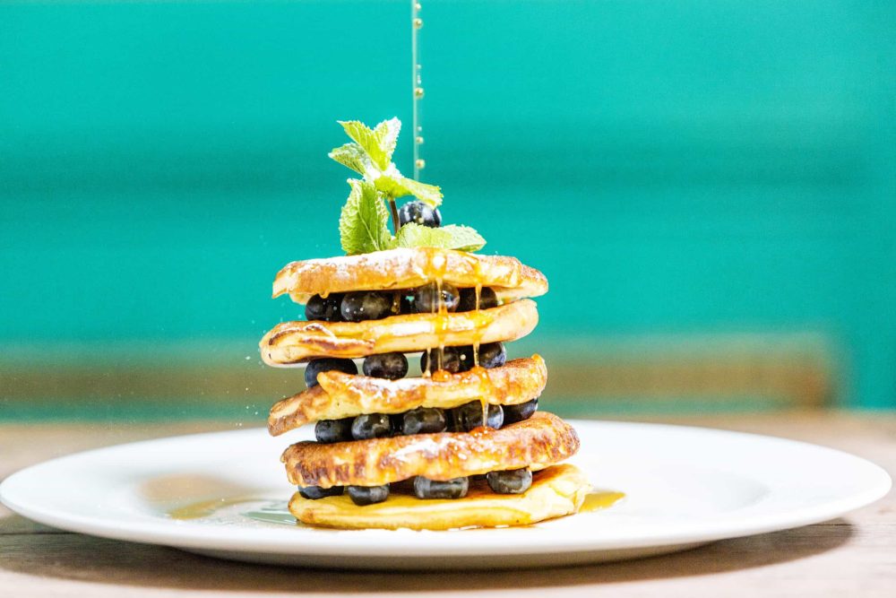 Fresh blueberry-topped waffle stack drizzled with honey, garnished with mint leaves, served on a white plate against a vibrant green background at FiFi's Italian restaurant.